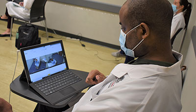 Nursing student sitting at a desk watching a patient on a laptop screen.
