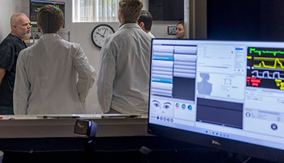 Looking though a window into a simulation room with a computer monitor displaying health data. 