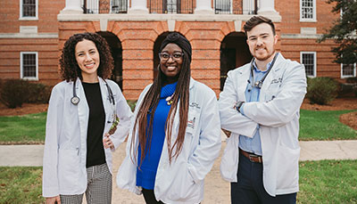 Three students wearing white coats standing in front of the School of Medicine building.
