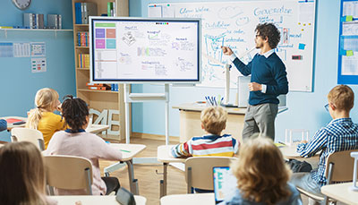Teacher in the front of a classroom of students pointing to a smart board. 