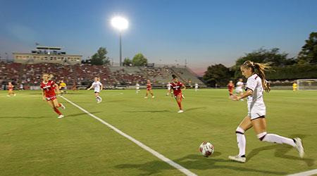 Women's soccer team playing on the field as the sun goes down behind them.