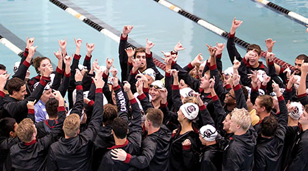 The swimming and diving team huddled together with their arms raised and cheering.
