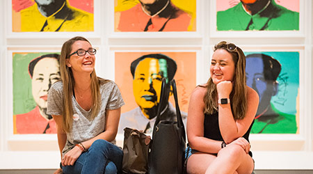 Two students sitting on a bench in a museum wiht pop art illustrations behind them. 