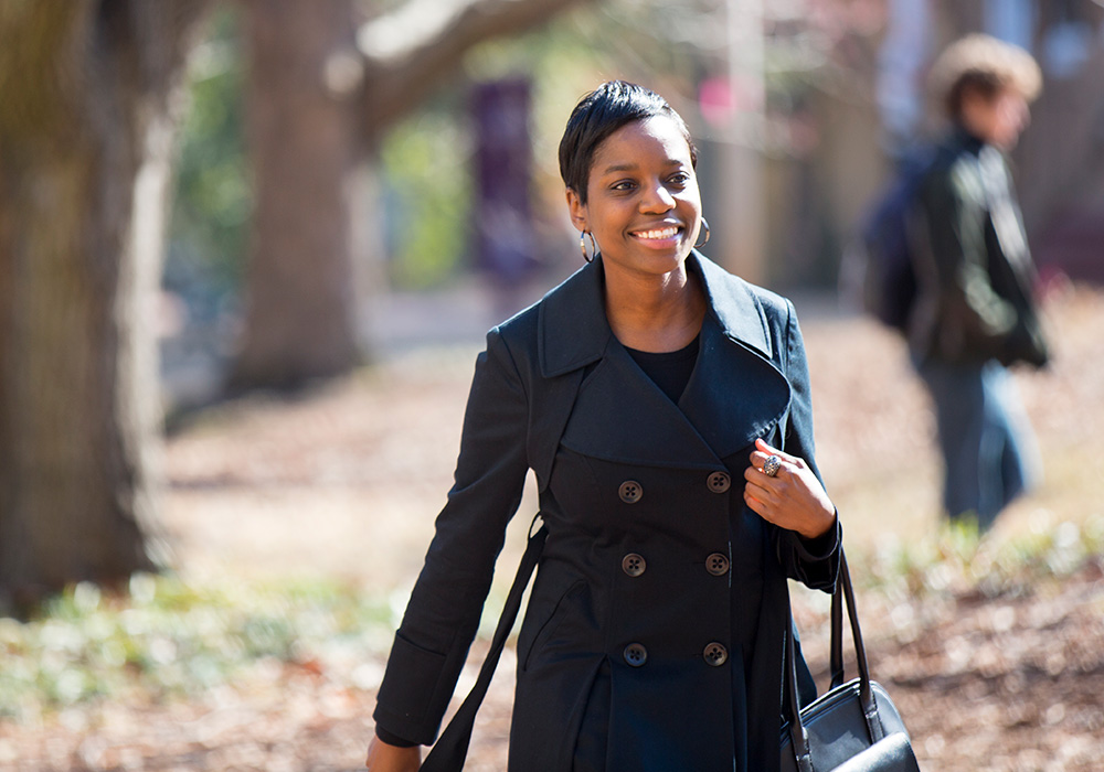 Young professional walking across campus outside wearing a coat and carrying a bag.