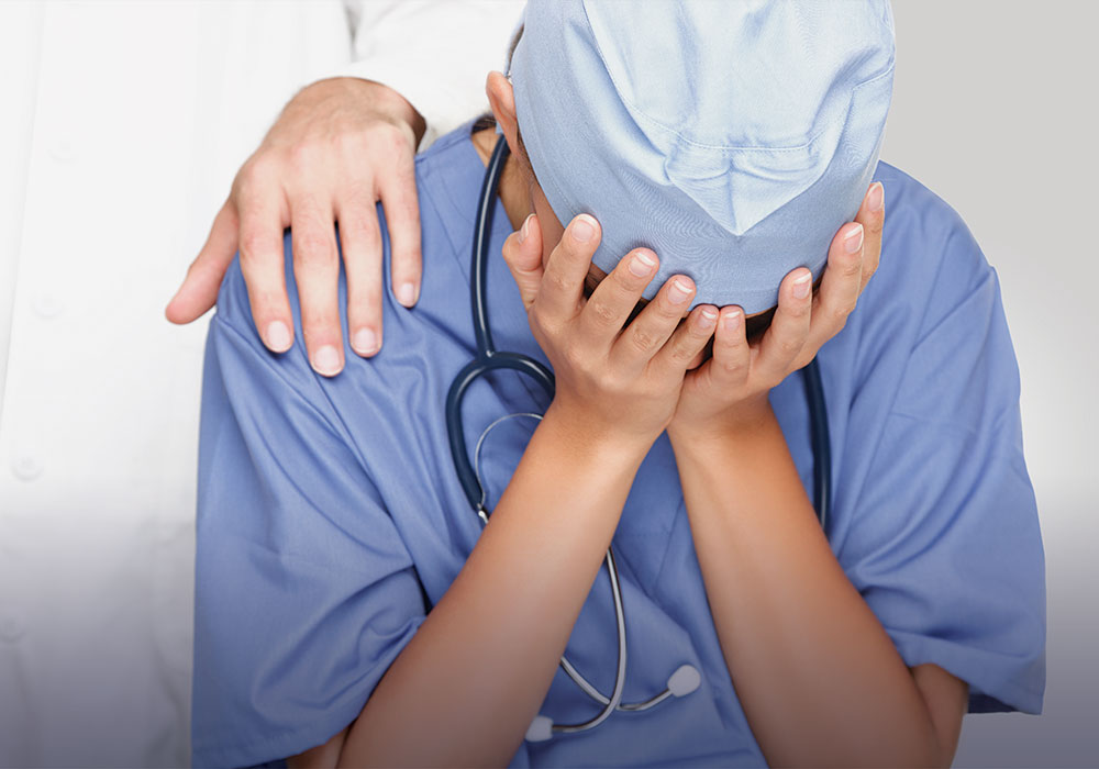 Person wearing scrubs with their head in their hands and a person consoling them with a hand on their shoulder. 