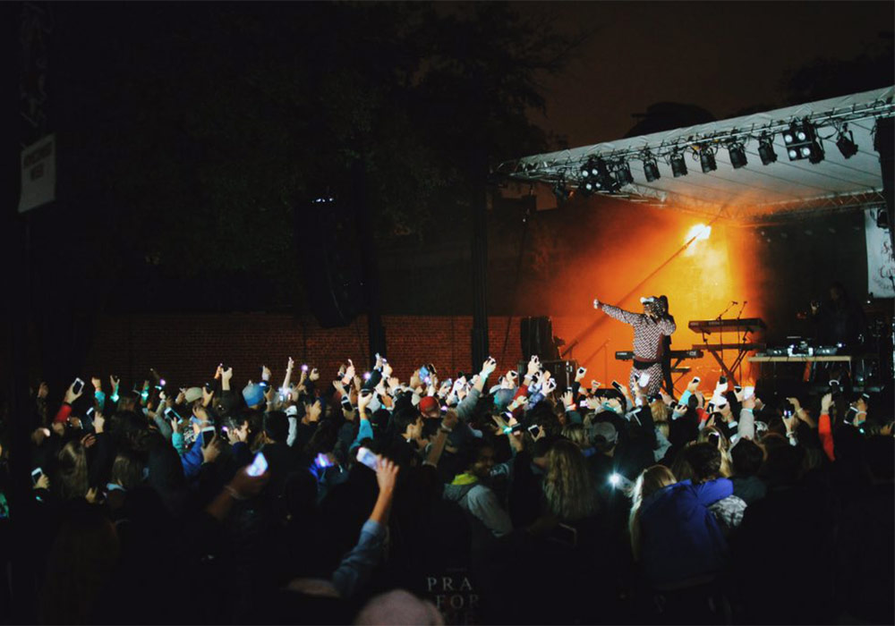 Crowd cheering in front of a stage with musician at night with glowing stage lights. 
