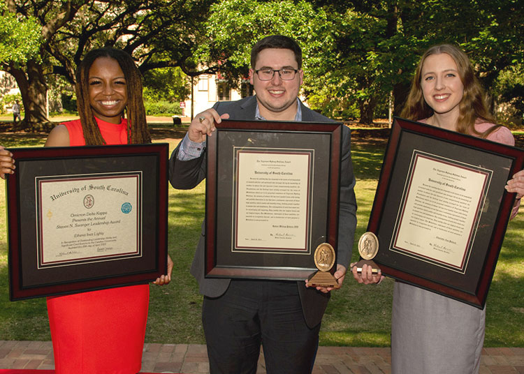 three students stand on the horseshoe holding large framed awards