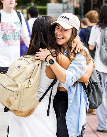 Two students hugging while smiling when running into each other on a campus sidewalk.