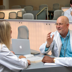 Medical students in white coats talk with a professor in a classroom setting during a teaching-hospital training session.
