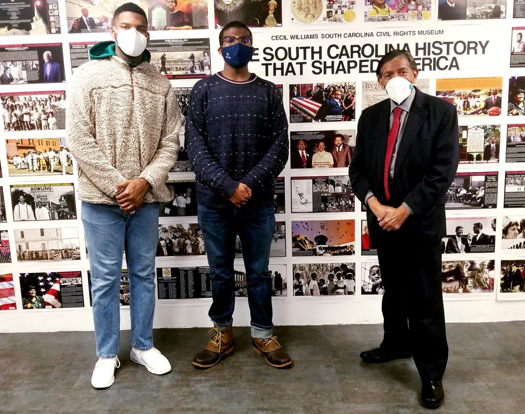 Three men, including Cecil Williams, pose for a photo in front of a collage wall of photos, with words that say “The South Carolina History that Shaped America” and “Cecil Williams South Carolina Civil Rights Museum.”