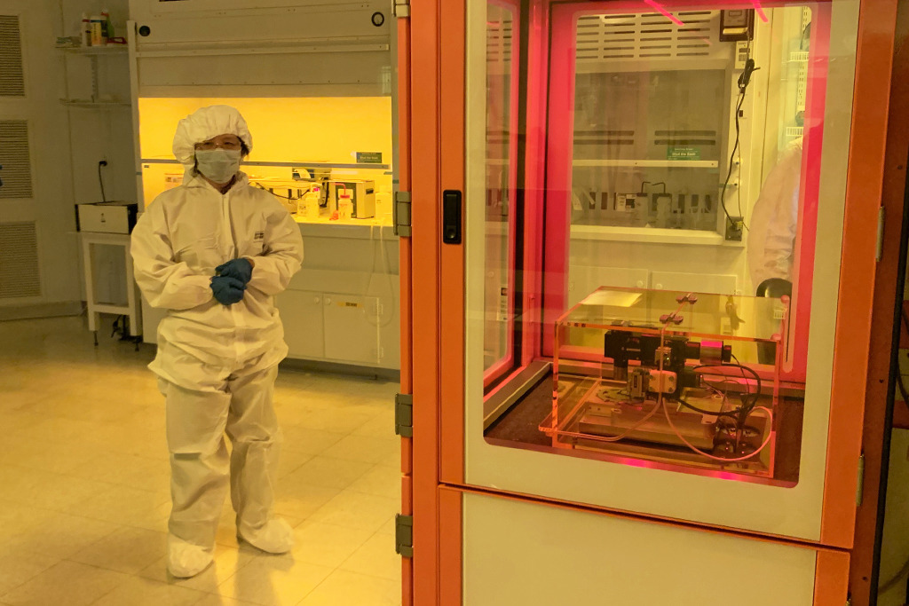 A woman dressed head to toe in white personal protective equipment stands slightly behind an orange and glass case containing a high tech piece of equipment called a Nanofrazor Explore. The light in the room has a yellow glow.