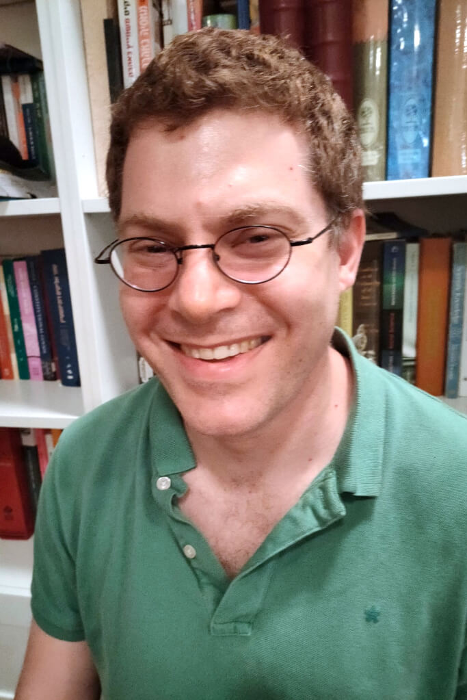 Smiling man with brown hair, green polo shirt, and glasses in front of bookshelf