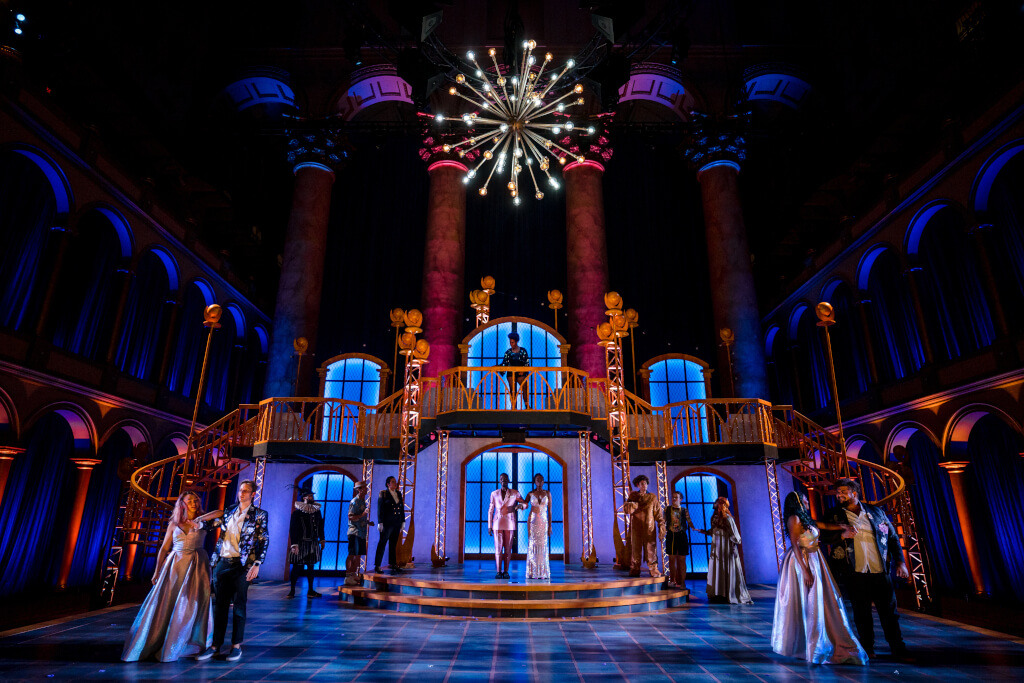 Wide angle shot of stage with colorful lights and thirteen actors in costume rehearsing scene