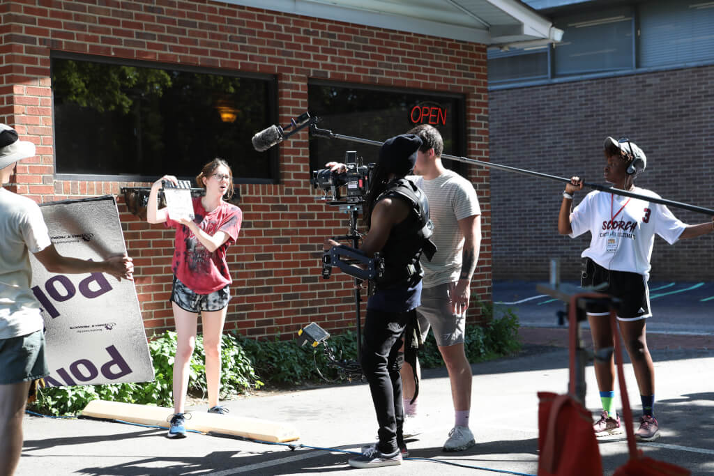 5 people gathered at a film set near a restaurant. There is a camera on a tripod, someone holds a long microphone and one person holds a clapperboard.