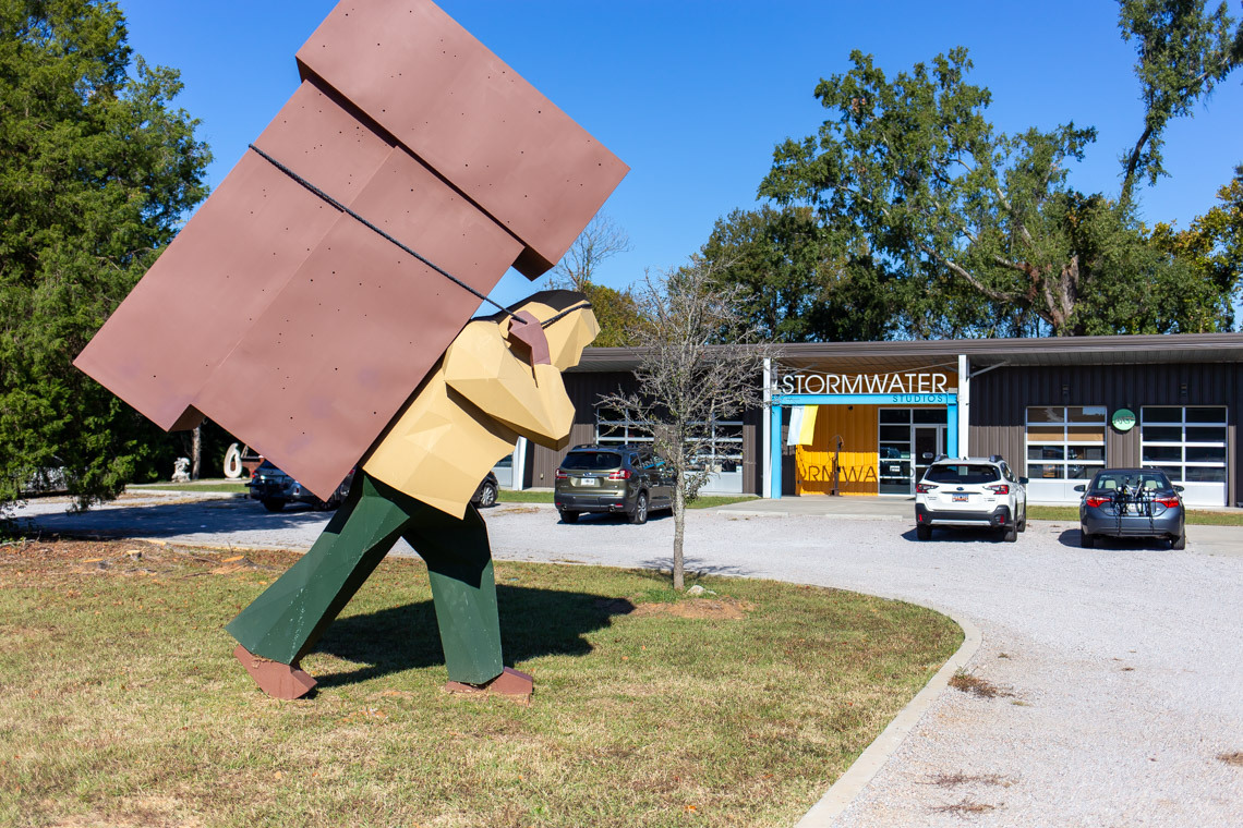 A sculpture on the front lawn at Stormwater Studios shows a human-type figure carrying a large block-shaped object on its back.