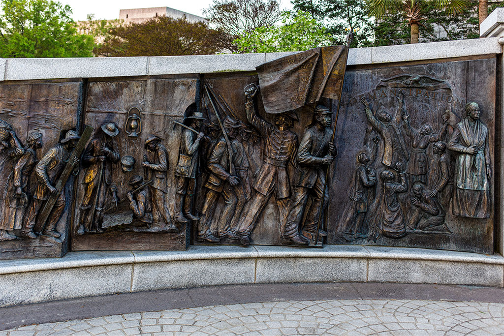 Bronze panels from the African American History monument at the South Carolina State House. This section shows a progression from enslaved individuals to soldiers marching with a flag to liberated people celebrating.