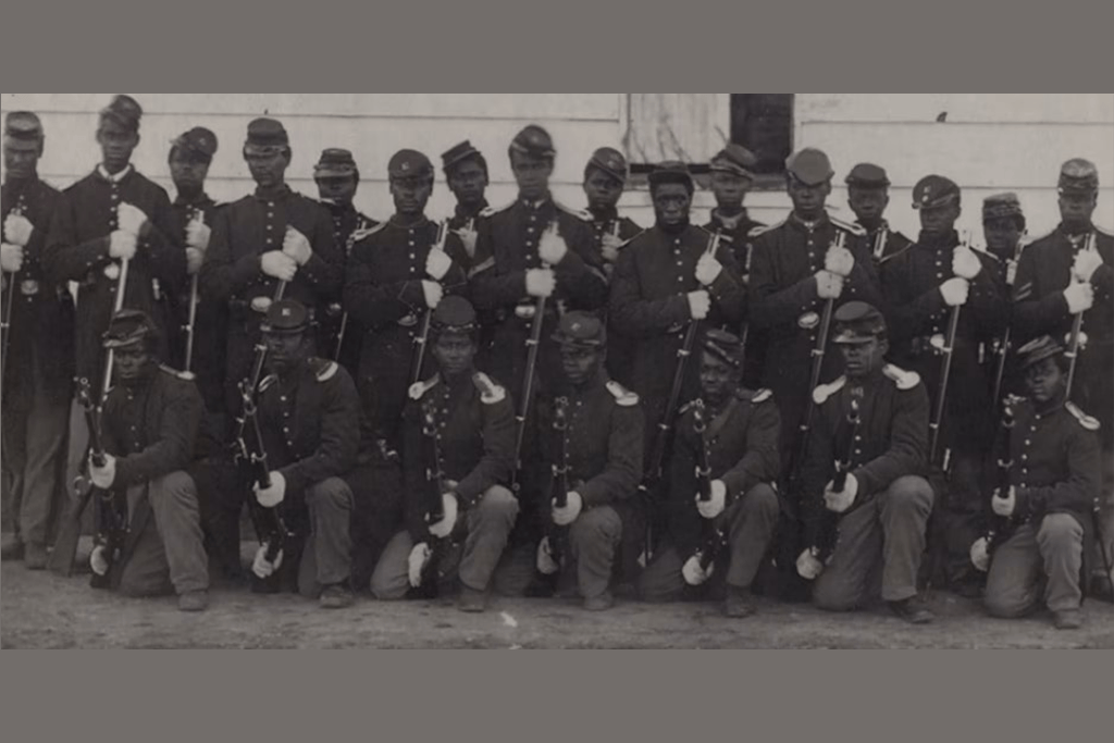 Black and white photo of Civil War Union troops. Rows of Black soldiers in Union uniform, one standing and one kneeling, with equipment.