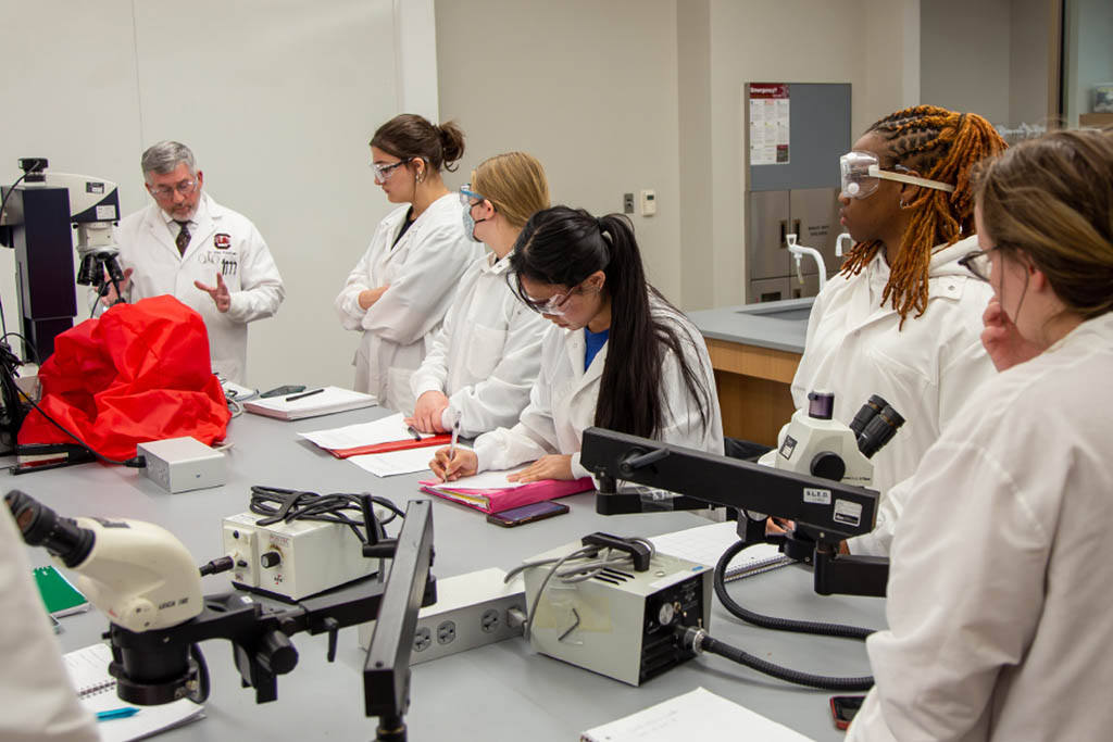 Students in white lab coats in the forensic chemistry lab taking notes.