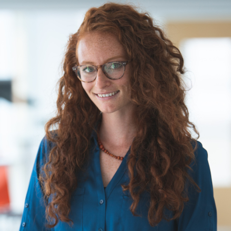 Nina Schreiner has fair skin with long, curly red hair and freckles. She wears tortoise shell glasses, a beaded necklace and a blue button down top while smiling at the camera.