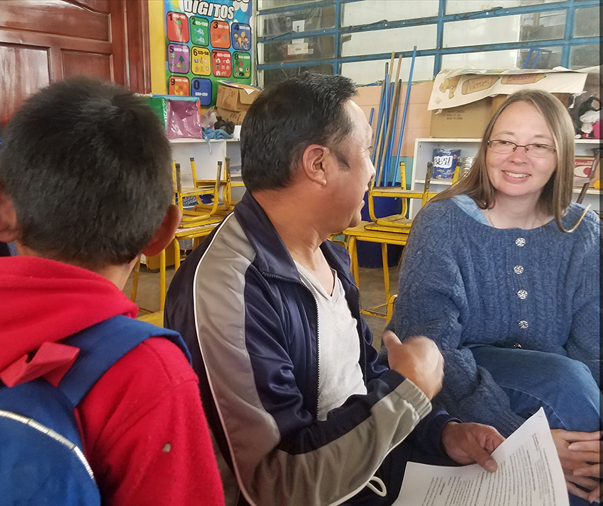 A researcher speaks with a teacher during a classroom interview while students sit nearby.