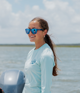 Lilly conducting research on boat with water and marsh in background.