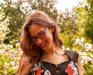 Bri Ash, a white woman with long brown hair, smiles in front of greenery