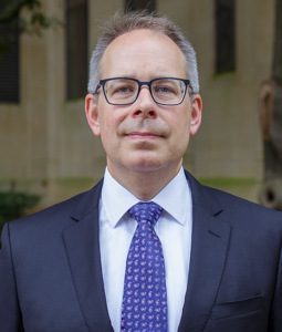 Carl Dahlman has short gray hair and black framed glasses, and wears a navy blazer, white shirt, and patterned blue tie.