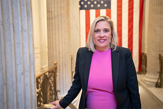woman in blue blazer with pink top on smiling in front of an american flag