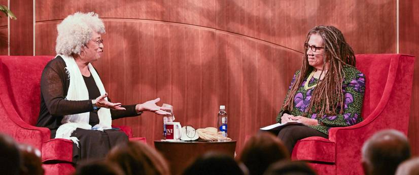 Angela Davis and Nikki Finney sit in comfortable red chairs on a presentation stage, facing each other while engaged in animated conversation.