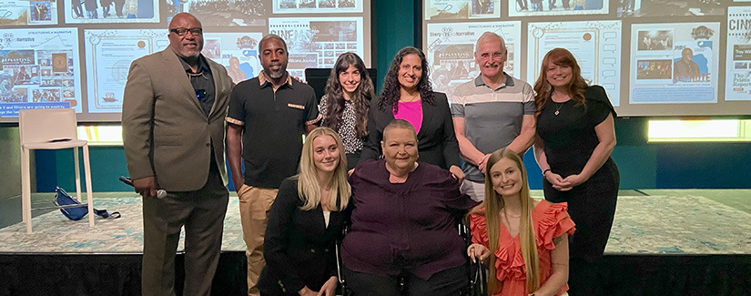 Group of nine people, including USC criminology Ph.D. student Christina Plakas, posing together on stage in front of a large presentation screen featuring prison advocacy and storytelling projects.