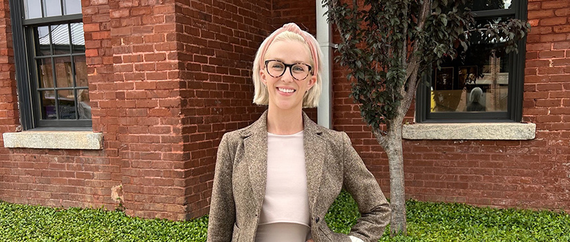 Elizabeth Connors wearing business attire with short blonde hair. She is standing outside by a brick building and bushes.