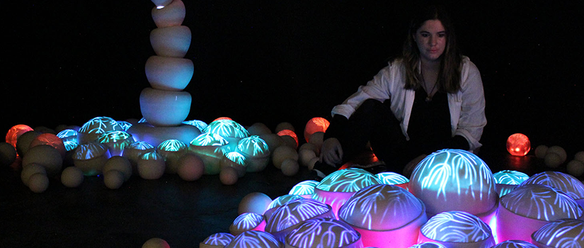 A person sits among Aimee Marcinko’s porcelain sculptures, glowing blue, purple and red with branching patterns beneath a tall, curved ceramic form.