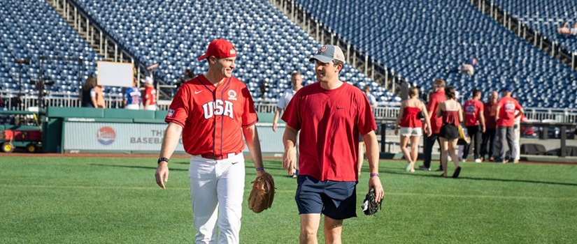 Two men walk across a baseball field, one wearing a USA baseball uniform and the other a red T-shirt and baseball cap.