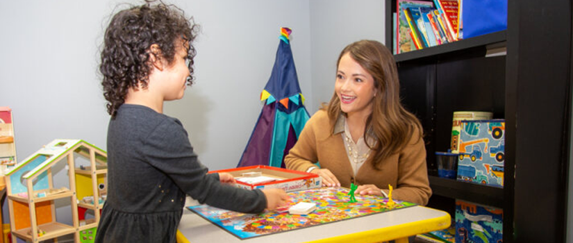 A young child plays a board game with a school counselor in a brightly decorated classroom filled with toys and books.