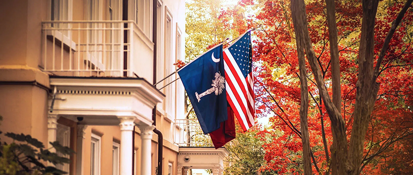 South Carolina and U.S. flags displayed outside a historic campus building framed by autumn trees on the University of South Carolina campus