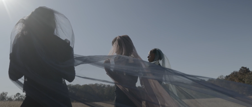 Three performers move through an open field draped in flowing translucent fabric, backlit by sunlight, with fabric trailing in the wind to create a layered, ethereal effect.
