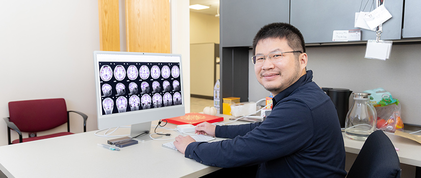 Assistant professor Feilong Ma sits at a desk in a psychology research lab, smiling at the camera, with a computer monitor displaying multiple brain scan images behind him.