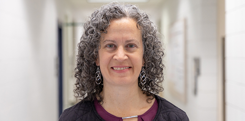 Caryn Outten, chemistry professor at the University of South Carolina, stands smiling in a campus hallway, wearing a dark jacket over a maroon top, with laboratory posters softly blurred in the background.