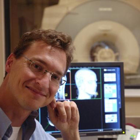 Chris Rorden sits in front of a computer with brain images. An MRI machine is in the background.