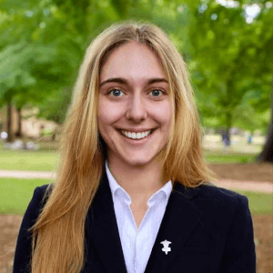Brynn has long blonde hair and smiles at the camera wearing a navy blazer while standing on the Horseshoe.