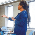 An African American professor teaches a class. They have long braids and are wearing a blue jacket.