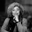 A black and white composite photograph shows Angela Davis from the waist up. She has shoulder-length curly hair and is wearing glasses, a long sleeve blouse, and a scarf.