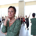 Anderson wears a green printed wrap dress and looks pensive standing in front of a mirror with the reflection of ballet students.
