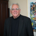 Knapp wears a black shirt and blazer and stands in an office with a large piece of abstact artwork.