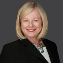 Randall wears a white blouse and black blazer with a turquoise necklace. She has a blonde bob and smiles at the camera.