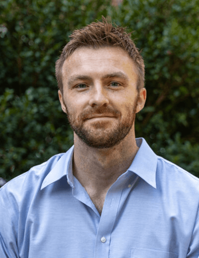 Alex Millen, new English faculty member at USC, wearing a light blue shirt and standing outdoors in front of greenery.