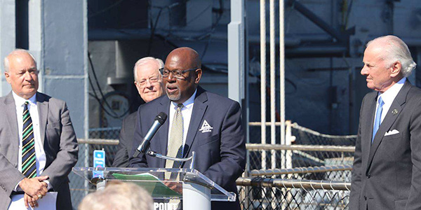 Ben Duncan speaks at a lectern beside a ship, with four other men standing nearby.