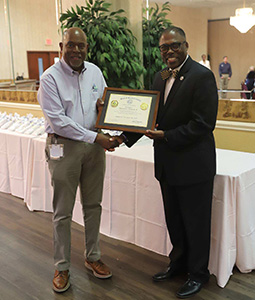 Ben Duncan and another man hold a framed certificate and shake hands at an indoor ceremony.