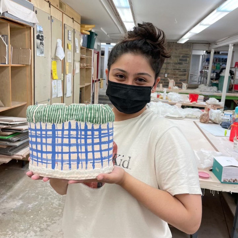 Catalina Marin holds a clay sculpture that she created at Penland over the summer. Catalina has olive-toned skin, dark brown hair and brown eyes. She is wearing a black face mask and white t-shirt.