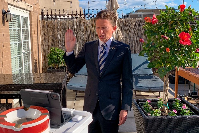 Geoff Palcher stands on a balcony. His right arm is raised as he is sworn in. He looks down at a table in front of him, where his laptop is pointed up at him.